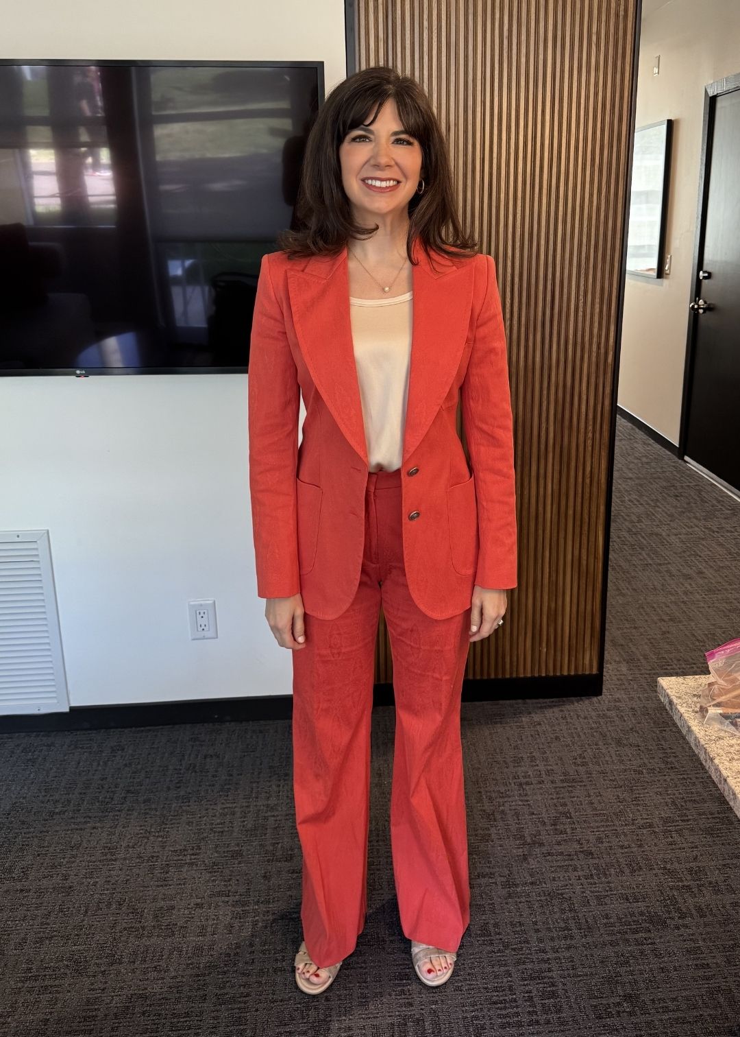 A woman stands indoors wearing a red suit with a beige top and open-toed shoes, smiling at the camera as if ready to share testimonials. She is positioned in front of a TV and wooden-panel wall.